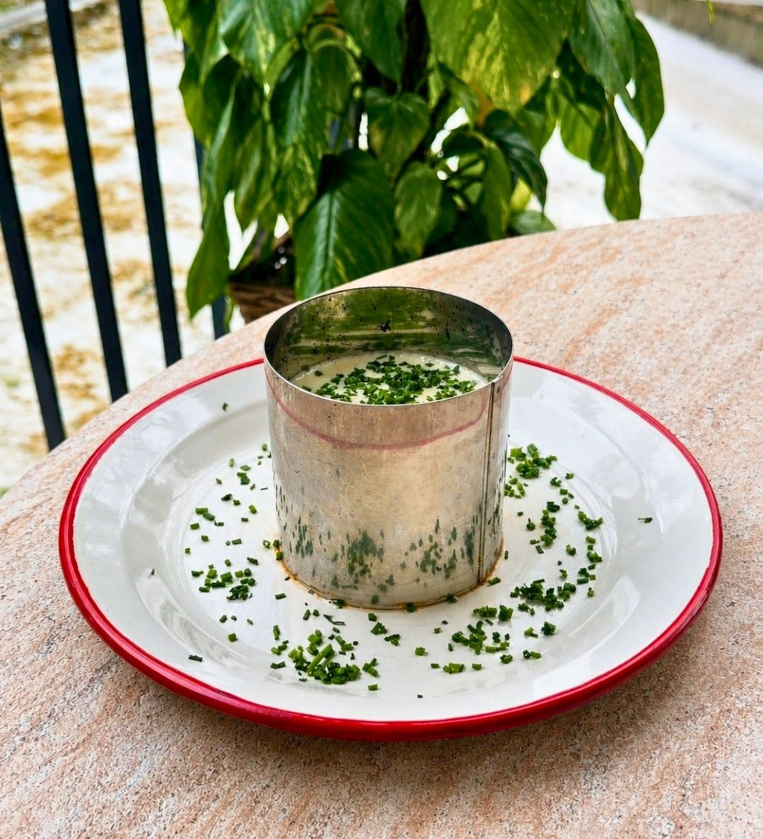 Xícara de metal com bebida de ervas verdes sobre um prato branco com borda vermelha em uma mesa ao ar livre.