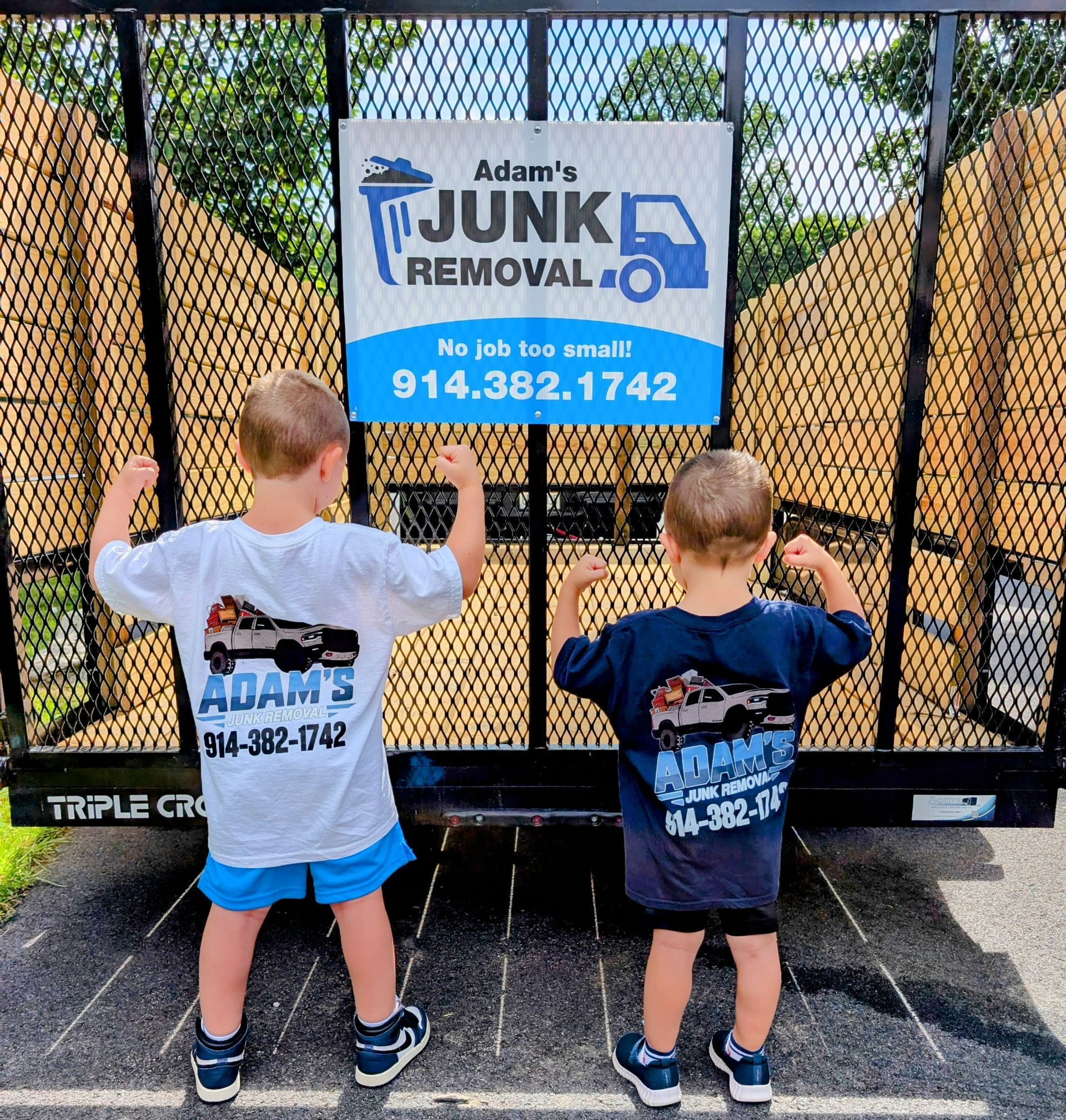 Two children flexing in front of an Adam's Junk Removal trailer, wearing matching shirts with the company logo.