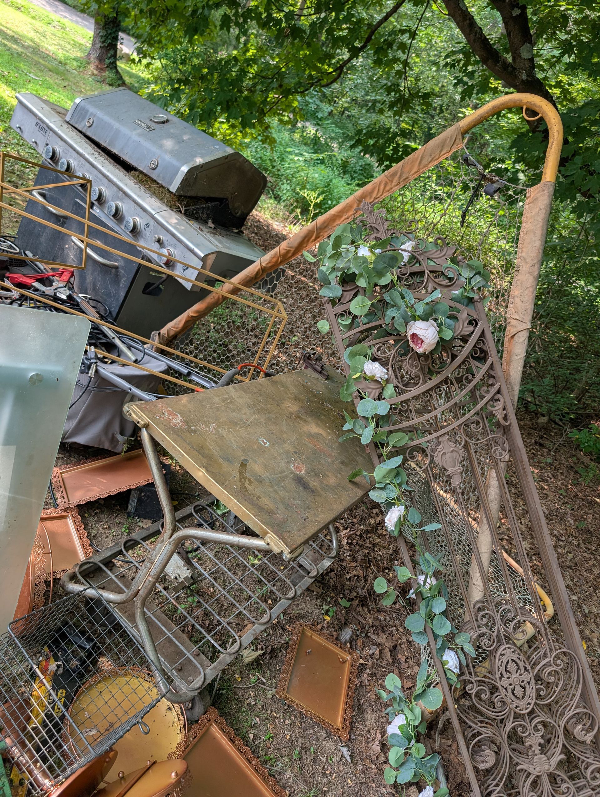 Pile of discarded items: metal chair, barbecue grill, rug, and climbing roses against a backdrop of green foliage.