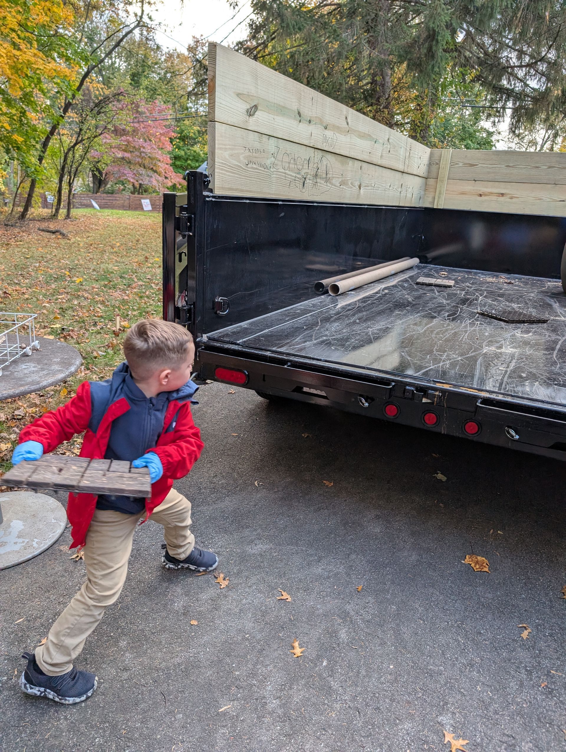 Child carrying a small wooden object towards a dump truck on an asphalt driveway.