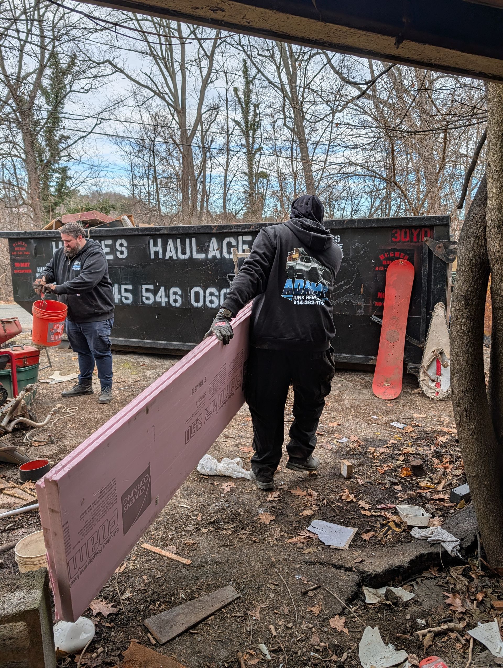 Two people by a dumpster, one carrying pink insulation, another holding a red bucket. Outdoor setting.