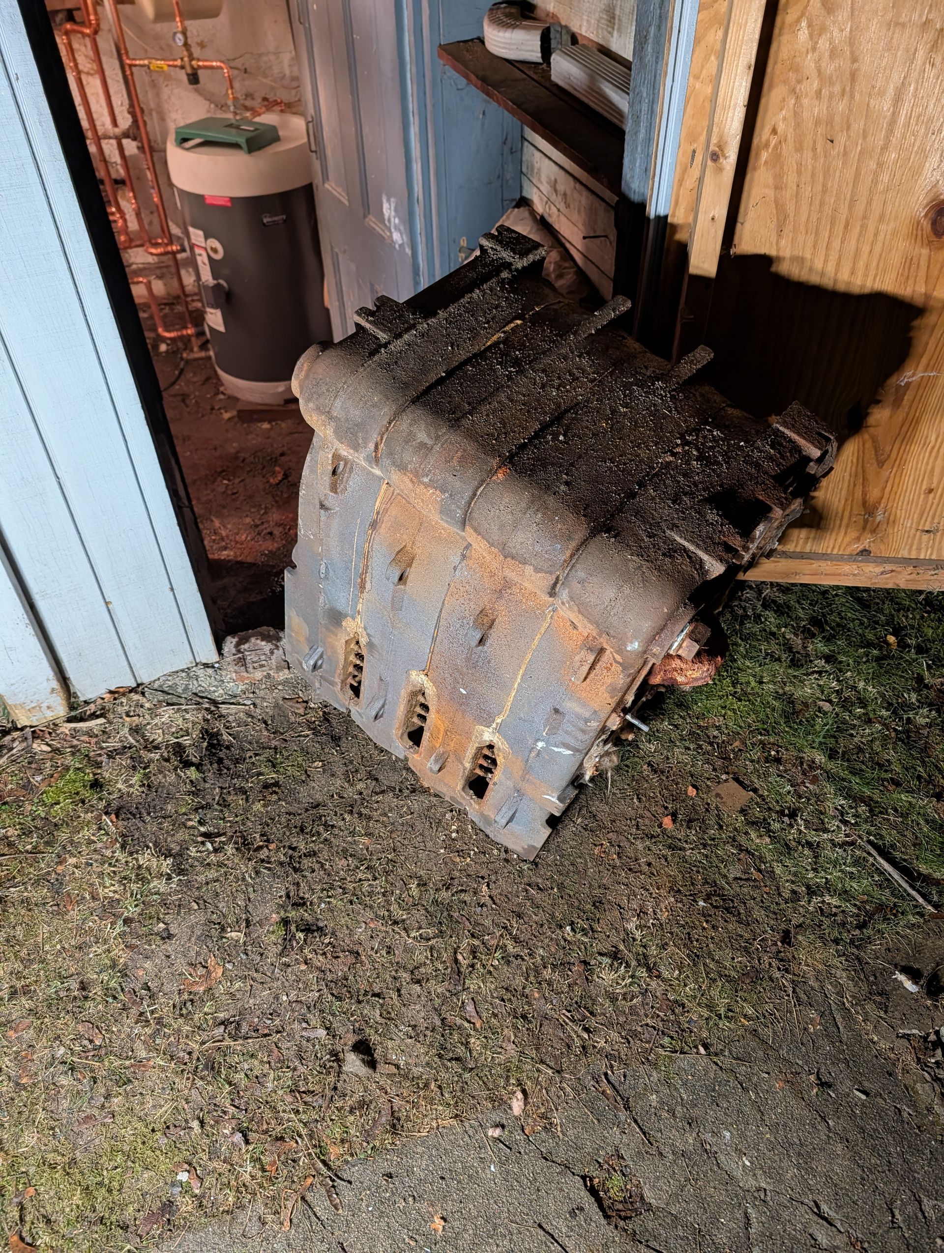 Old, rusty metal object leaning against a wall and wooden panel. Ground is covered in debris.