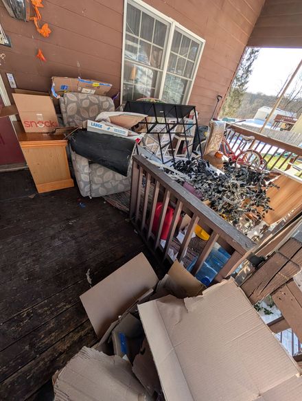 Porch cluttered with cardboard boxes, furniture, and debris near a window and railing.