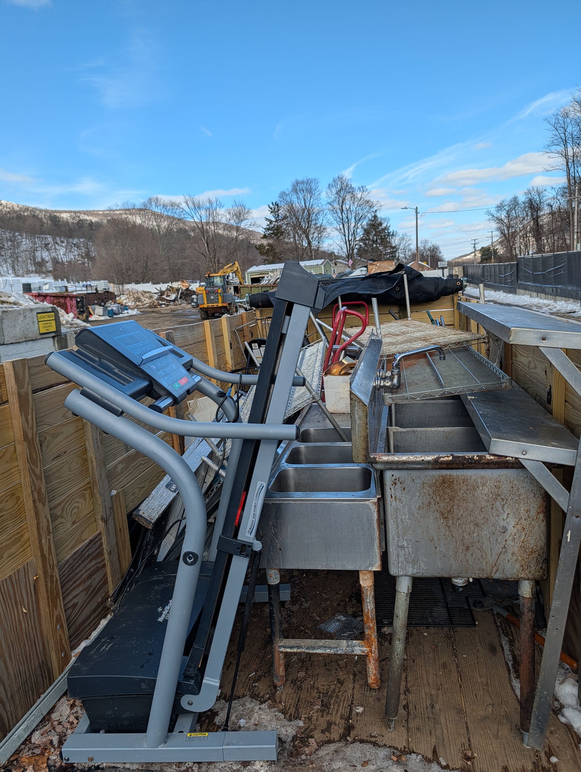 A treadmill and stainless steel sinks are piled in a debris-filled outdoor space, with snow on the ground.