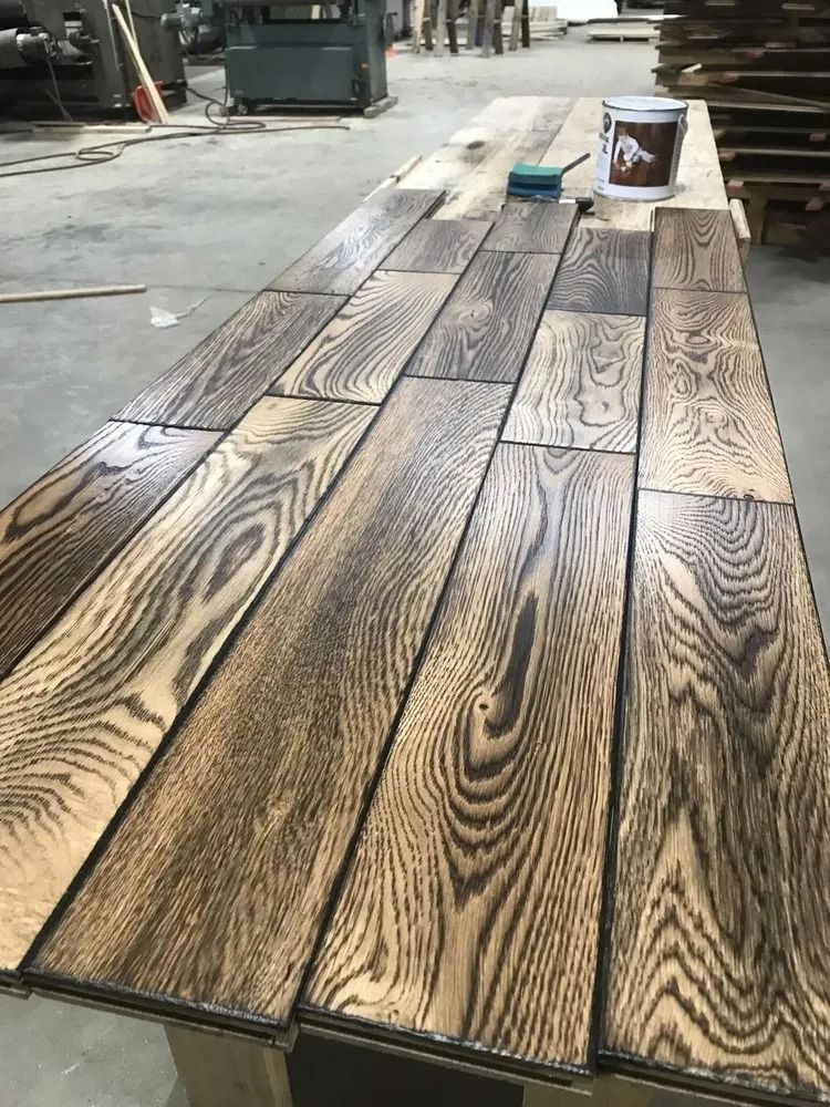 Stained wood planks laid out on a concrete floor with a paint bucket and brush nearby