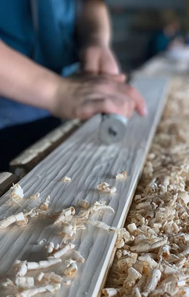 Hands planing a wooden board, with curls of shavings on a workbench.