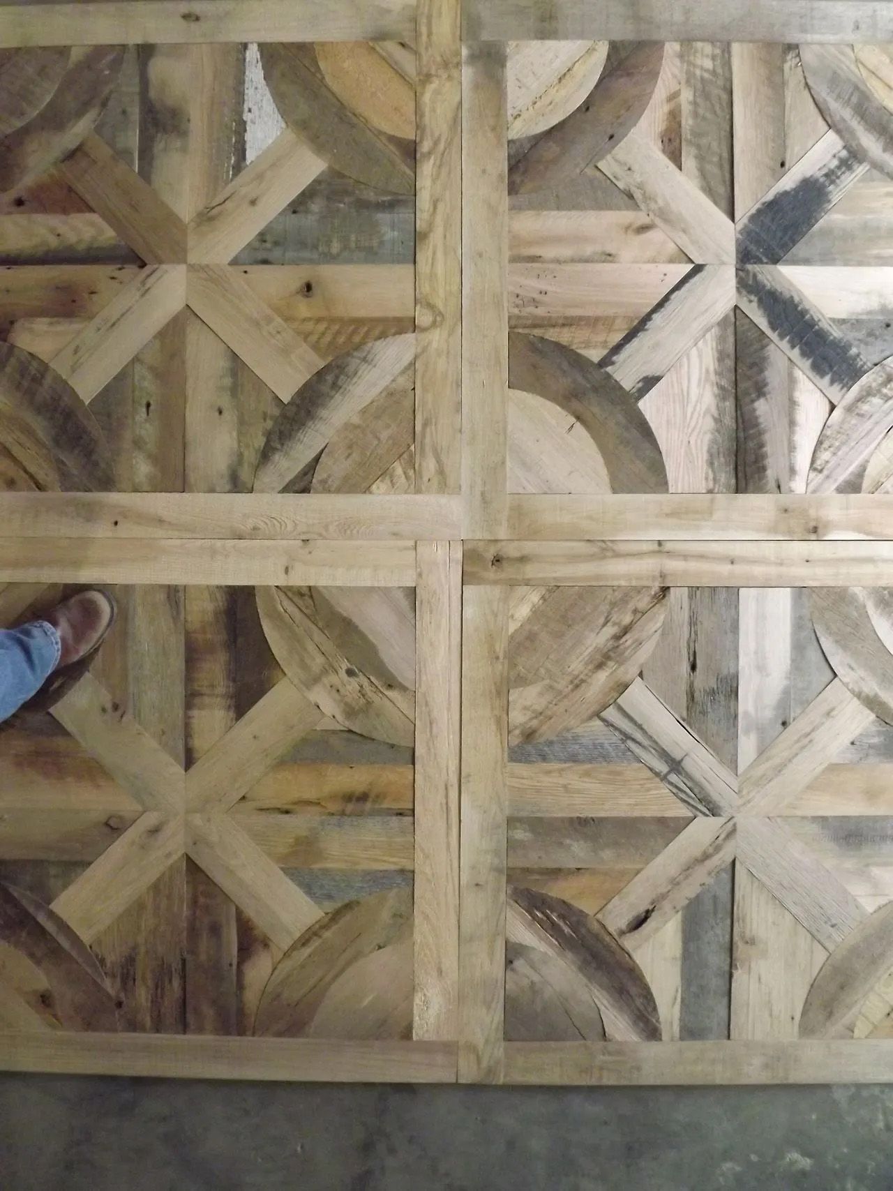 Wooden geometric tile pattern in beige and gray tones, with a person’s shoe visible at the lower left edge.