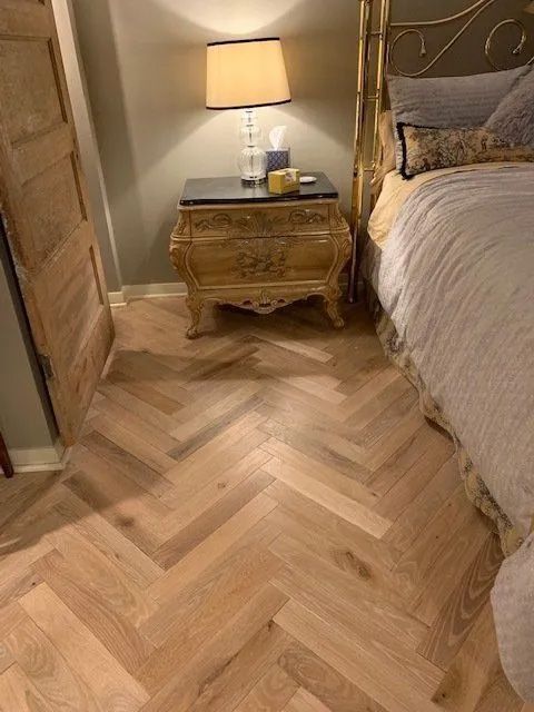 Bedroom corner with herringbone wood floor, upholstered bed, and lit bedside table lamp.