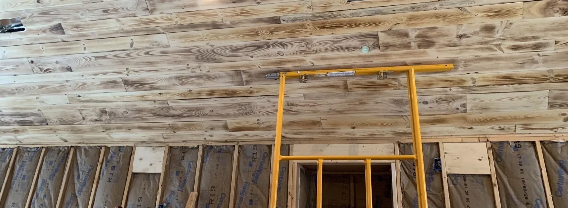 Rustic wooden wall with a yellow ladder leaning against it and barred window openings below.