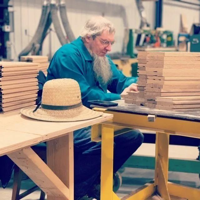 Worker in a teal shirt sits at a workbench with stacks of wooden boards and a straw hat nearby.