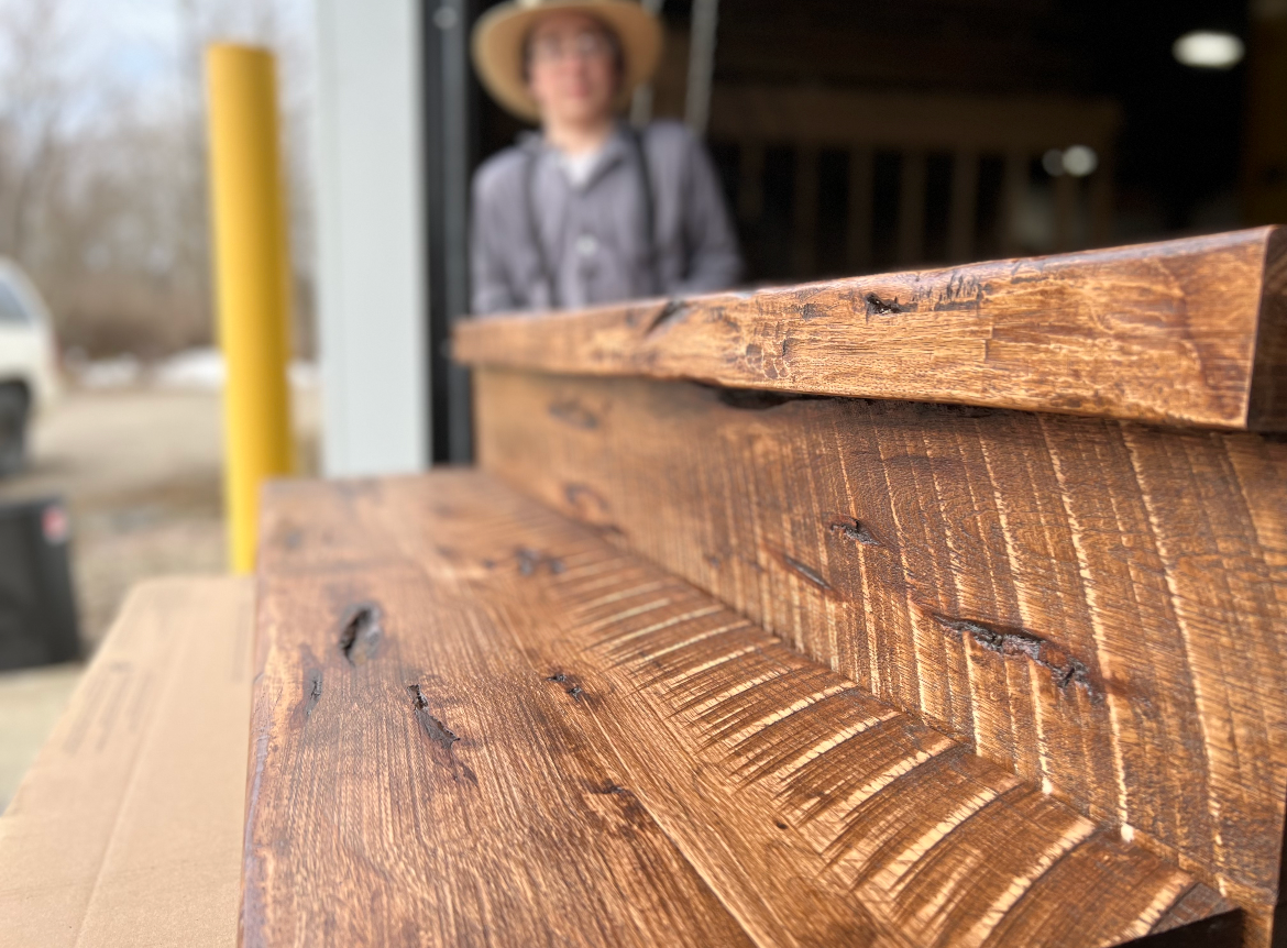 Close-up of stacked wooden beams in a workshop, with a blurred person standing in the background.