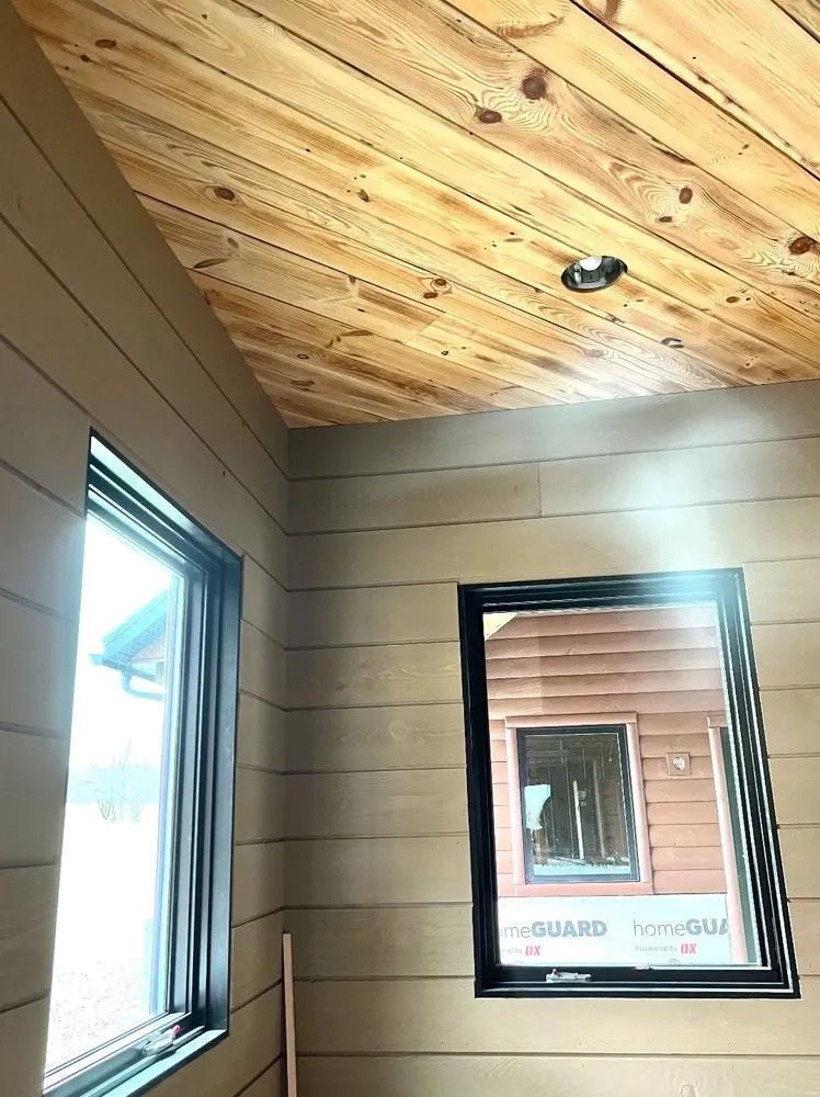Small room with wood ceiling, beige walls, and two black-framed windows.