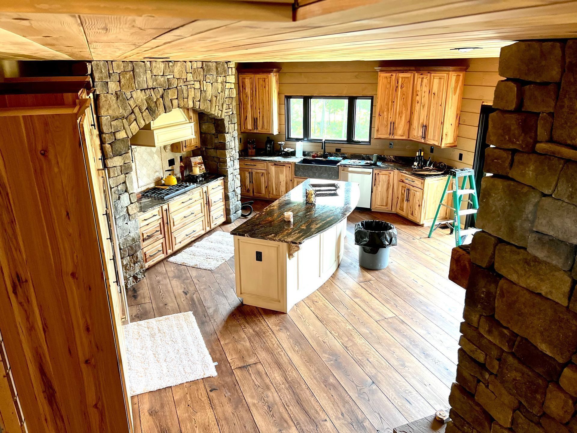 Rustic kitchen with stone fireplace, wood cabinets, island, and large windows on a wooden floor.