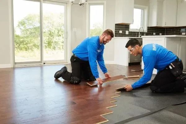 Two installers in blue shirts laying wood flooring in a bright room