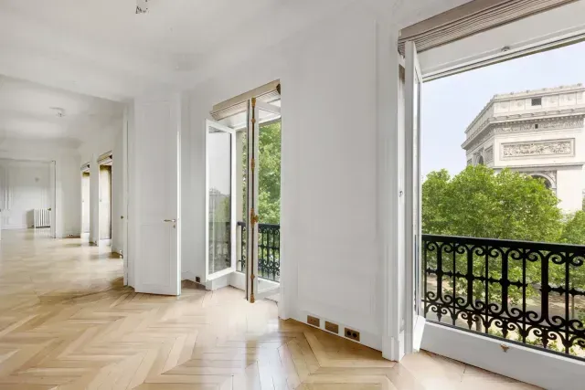 Bright empty room with herringbone wood floors, open balcony doors, and city view through large windows