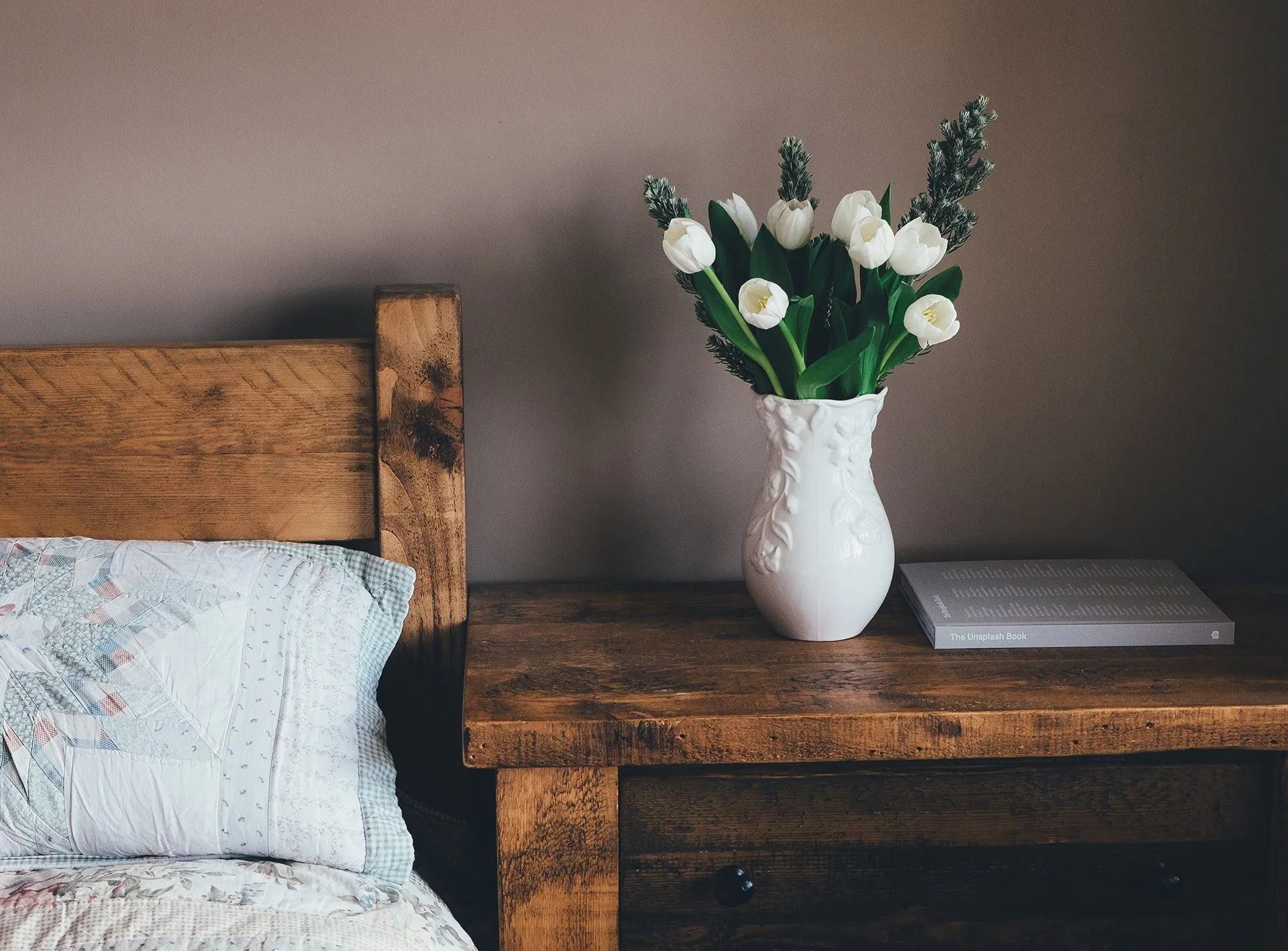 White tulips in a vase on a wooden bedside table beside a bed with blue bedding