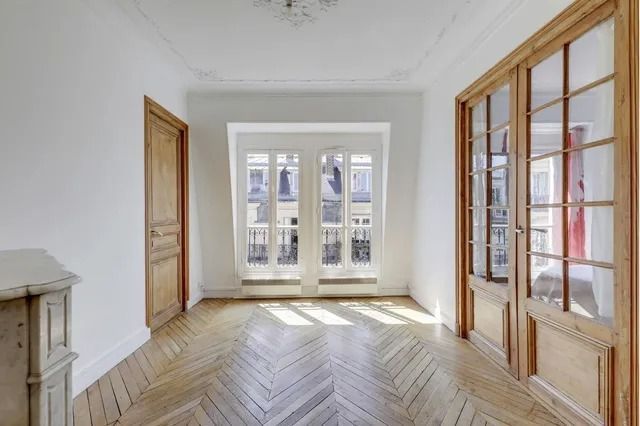 Bright empty room with herringbone wood floors, white walls, and tall windows letting in sunlight
