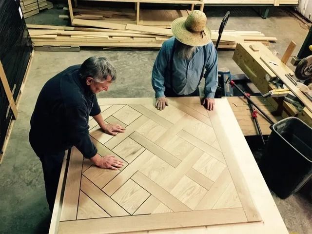 Two workers fitting a herringbone wood panel on a large table in a workshop