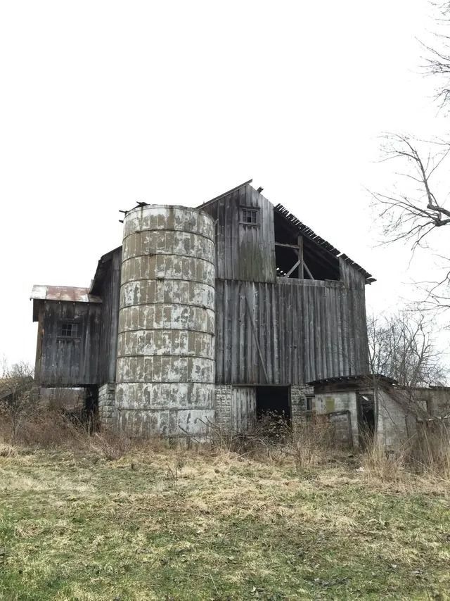 Old weathered barn with a cylindrical silo in an overgrown field under a gray sky