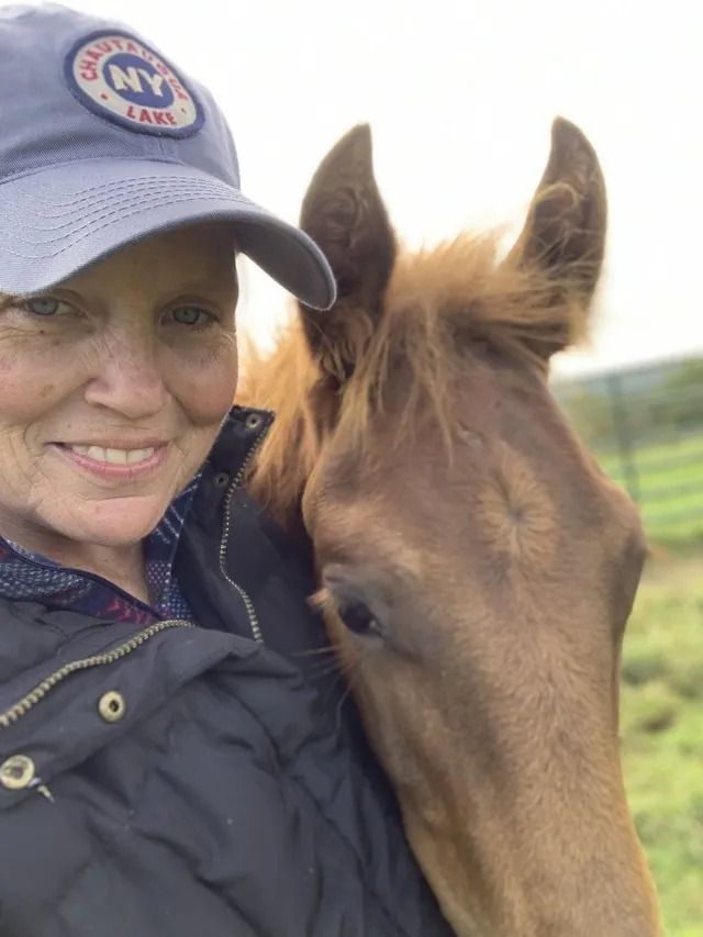 Woman in navy cap smiling beside a brown horse in a grassy field