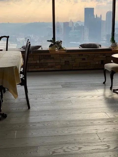 Sunlit dining room with wooden floor, table and chairs, overlooking a hazy city skyline through large windows