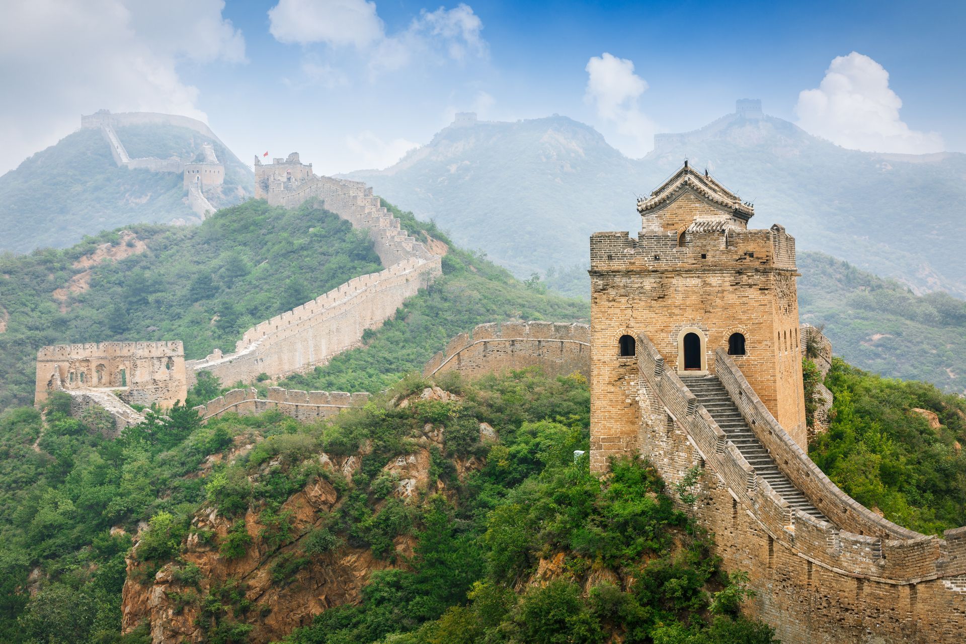 Great Wall of China winding over green mountains under a hazy sky, with a watchtower in the foreground