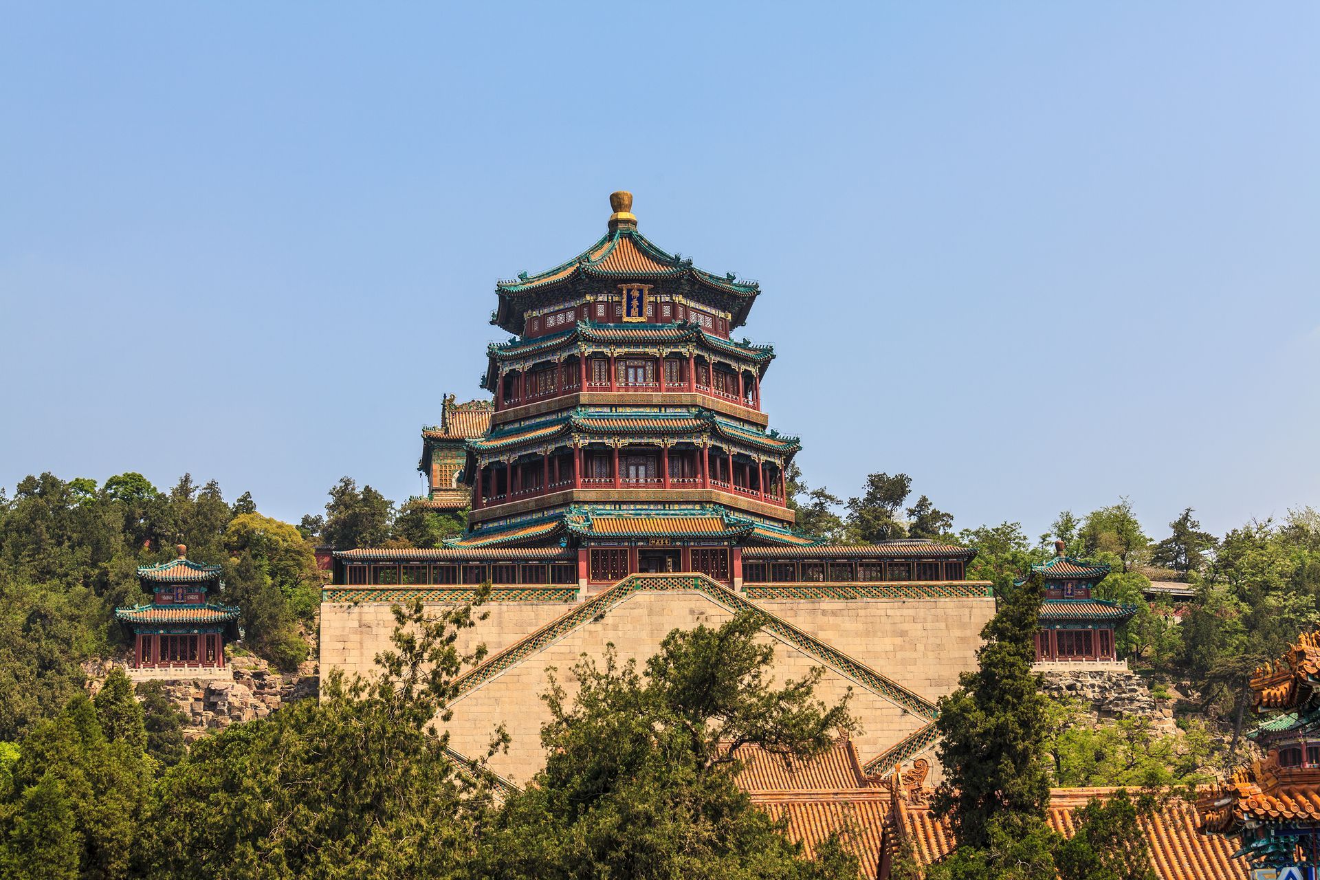 Multi-tiered traditional Chinese pavilion with red and green roofs above a stone wall and trees under a blue sky