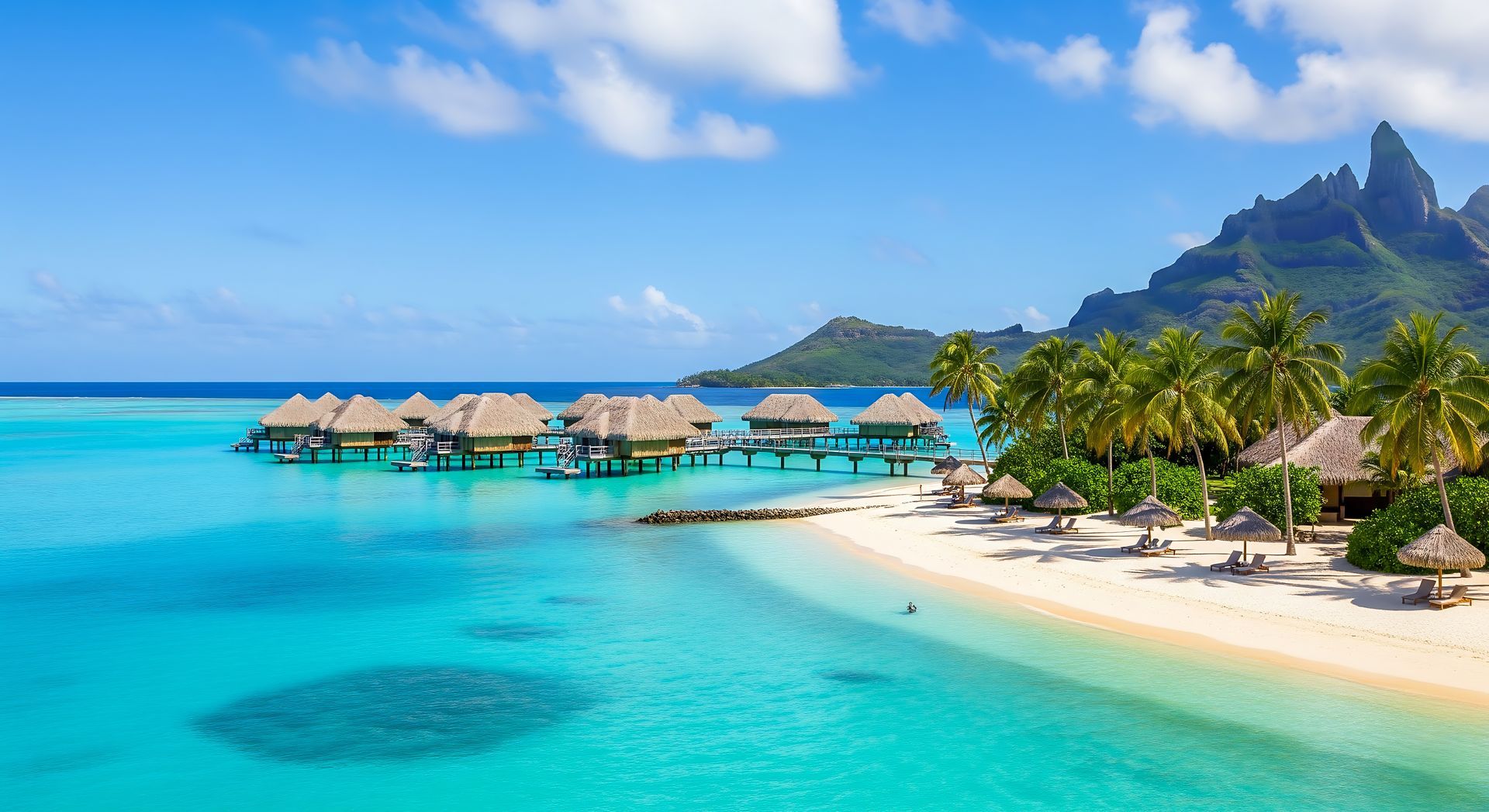 Tropical beach with turquoise water, overwater bungalows, palm trees, and mountains under a blue sky