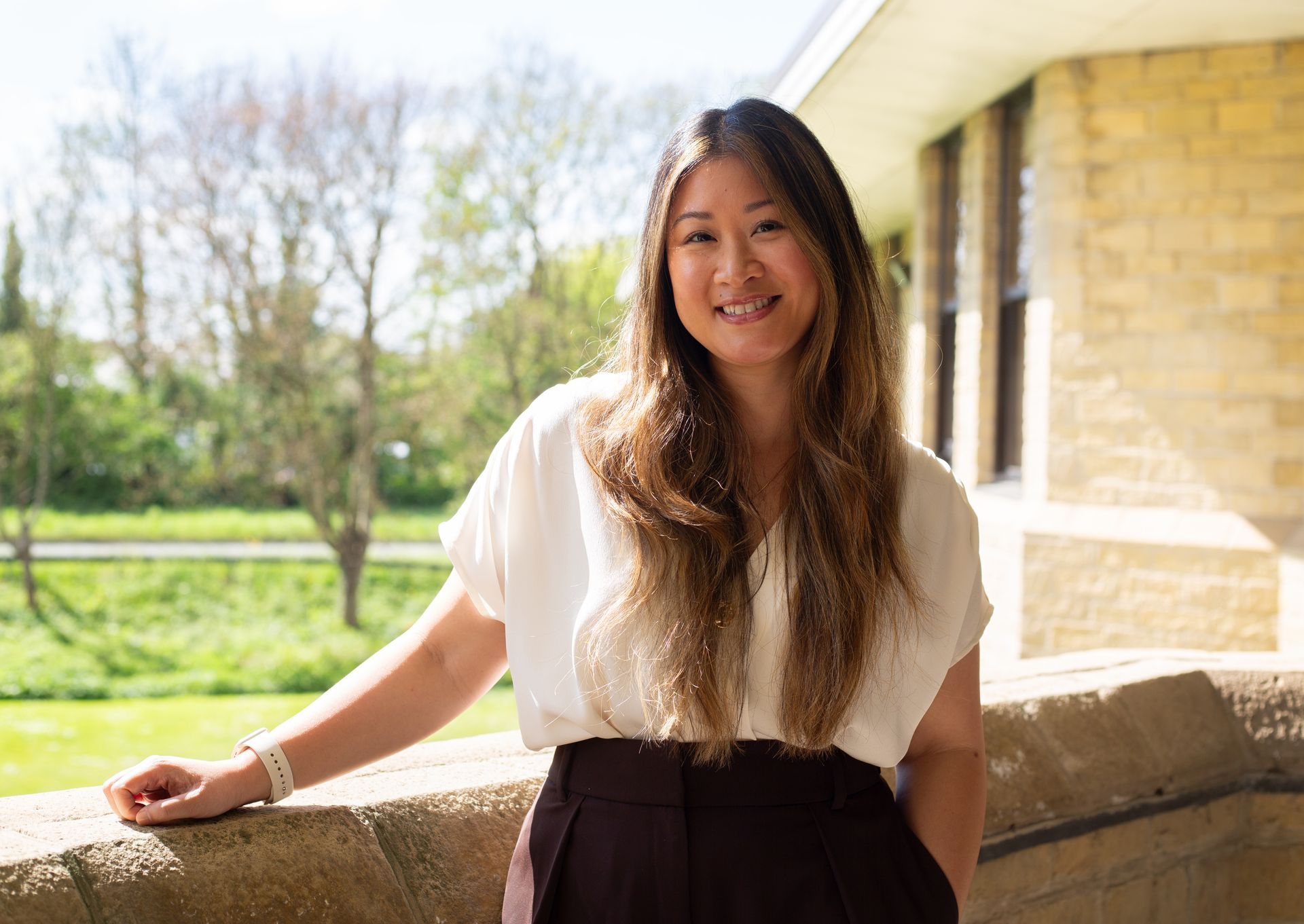 Smiling woman leaning on a stone railing outdoors beside a building in sunlight