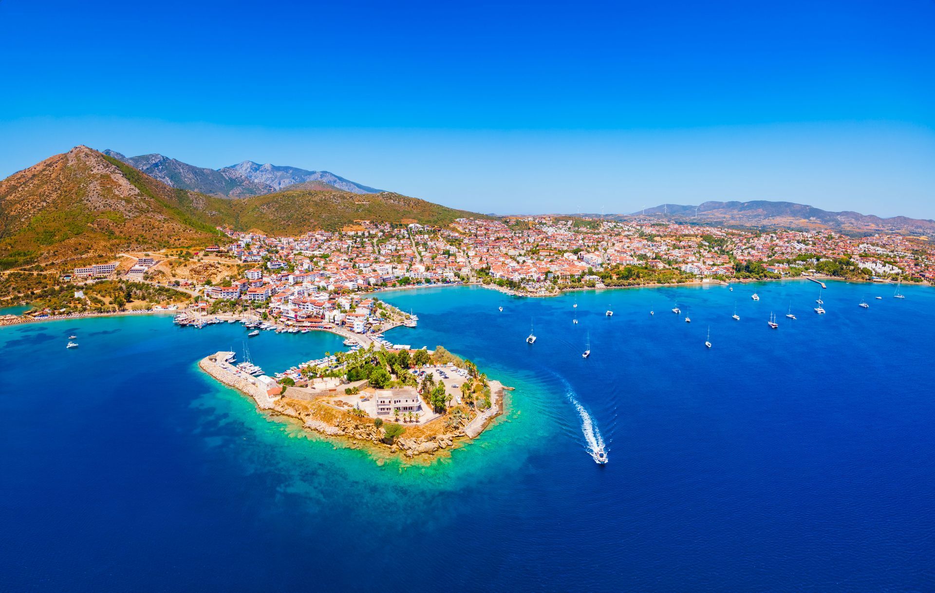 Aerial view of a coastal town on a turquoise bay with a small island and boats in deep blue water
