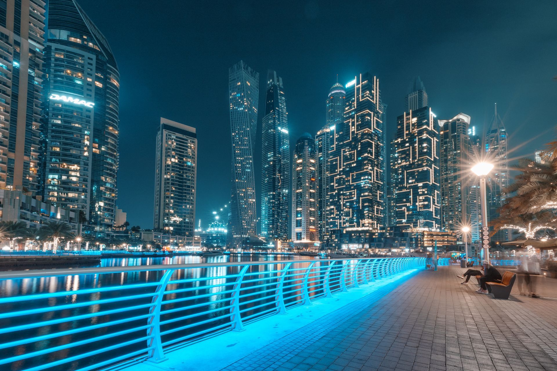 Nighttime city skyline with illuminated high-rise buildings and a blue-lit pedestrian promenade by the water