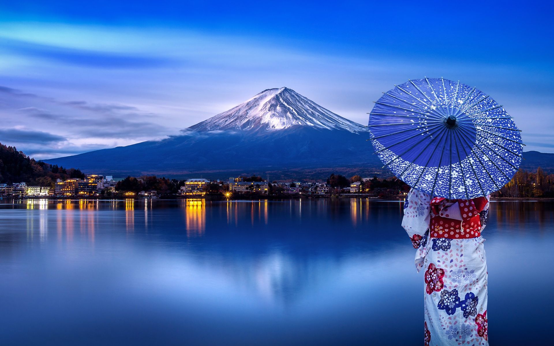 Snowy mountain reflected in a calm lake at dusk, with glowing lights and a blue parasol in the foreground