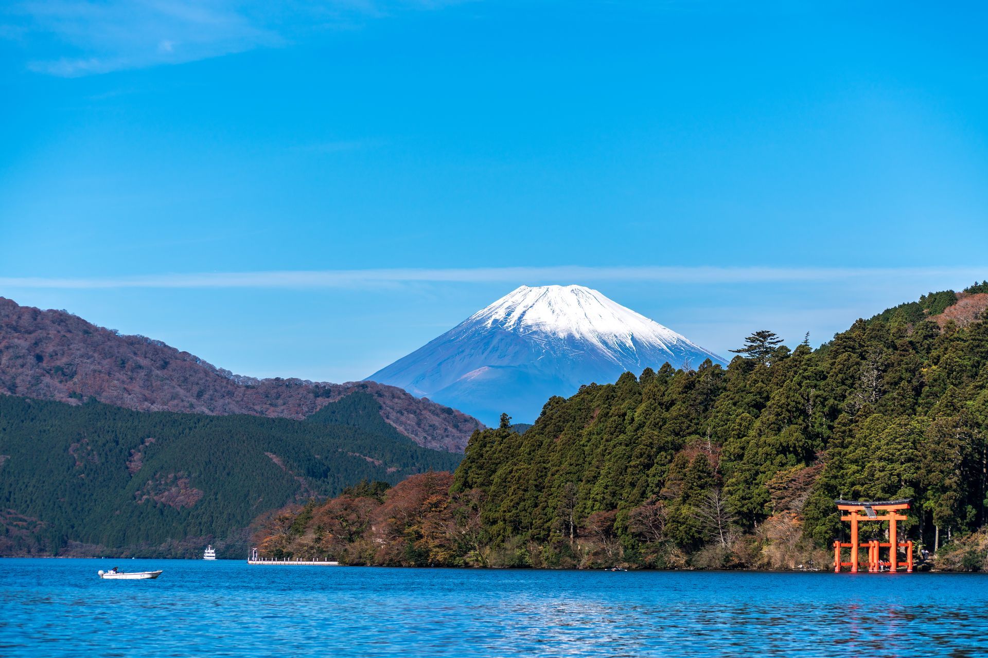 Snow-capped Mount Fuji over blue lake with forested hills and a red torii gate on the shore