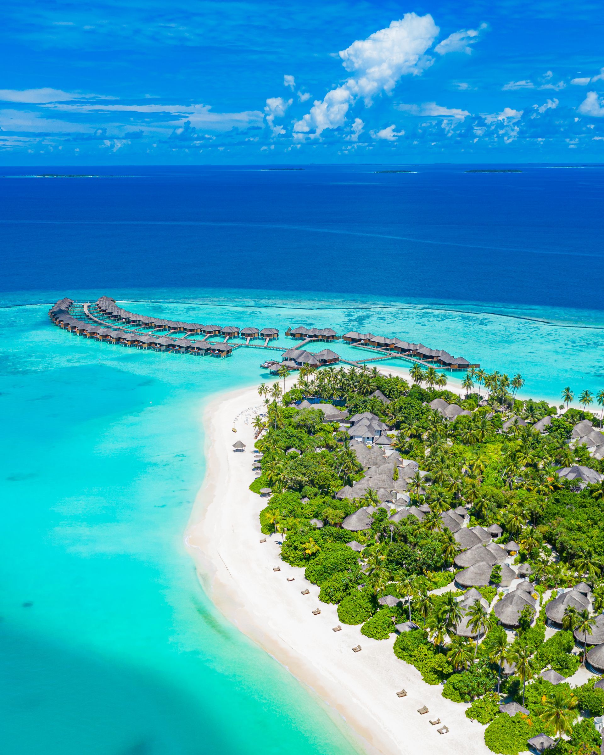 Aerial view of a tropical island with white sand, turquoise water, and overwater bungalows under a blue sky