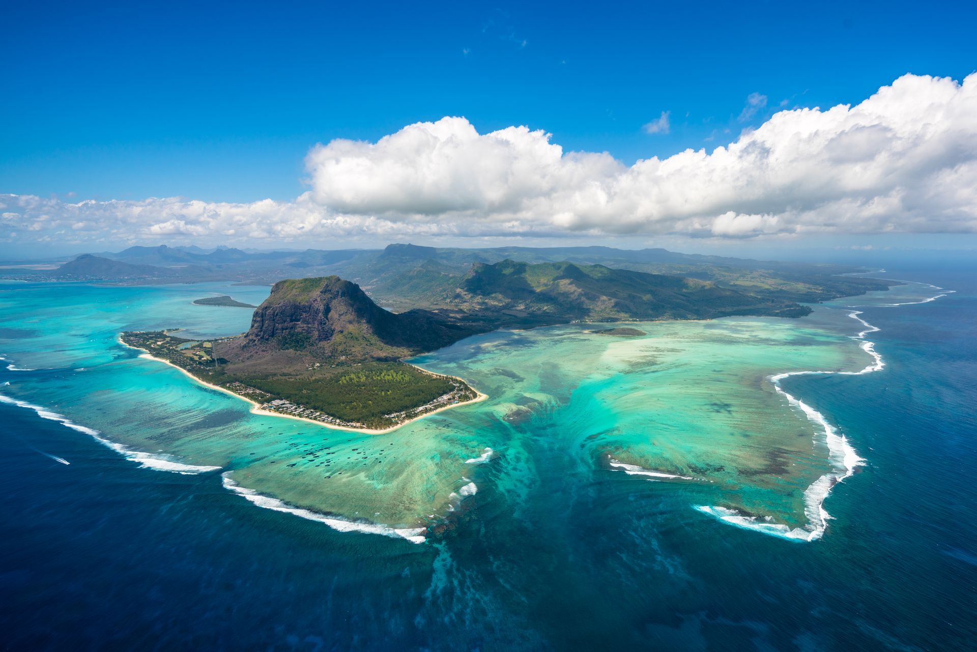 Aerial view of a tropical island with turquoise lagoons, white sand, and dark blue ocean under a bright sky