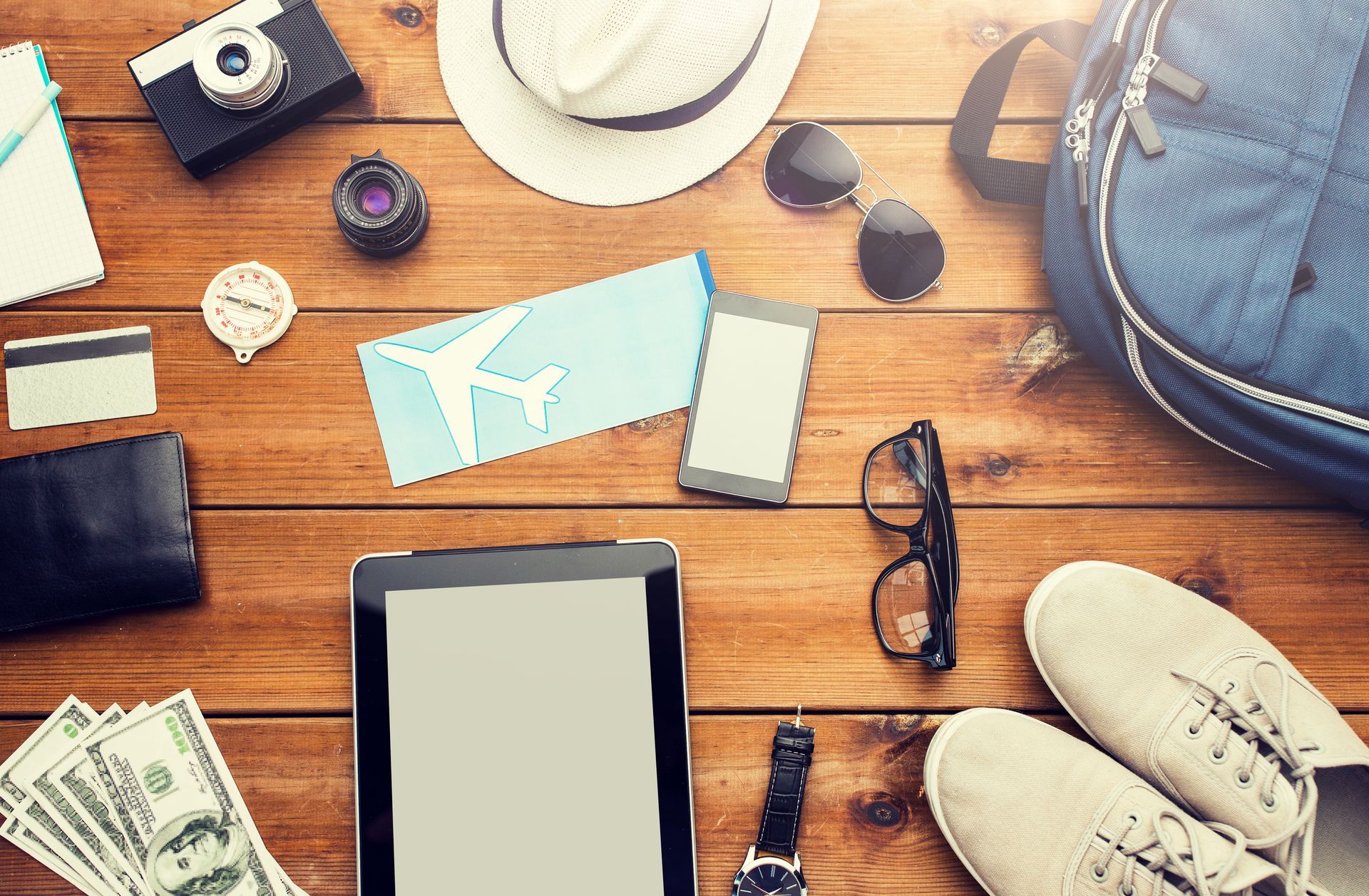 Travel essentials laid out on a wooden table: hat, tablet, camera, map, glasses, sneakers, cash, and backpack