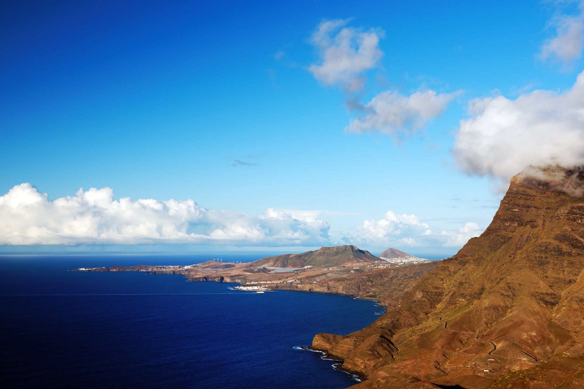 Coastal cliff overlooking deep blue ocean and distant town under a bright blue sky with clouds