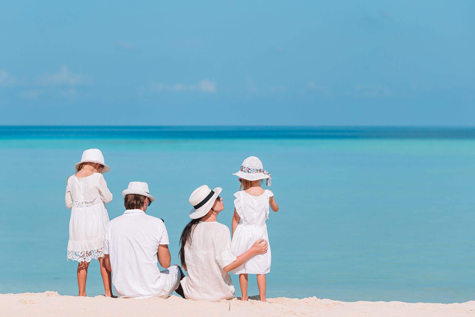 Four people in white sit and stand on a sandy beach facing calm turquoise water under a clear blue sky.