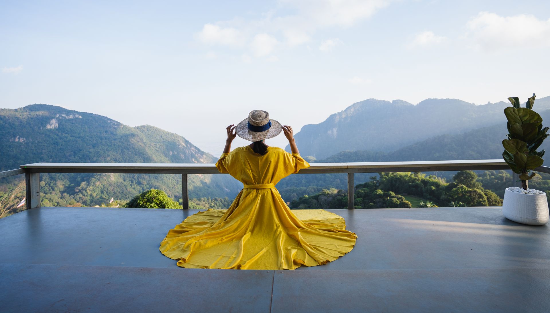 Person in a yellow dress standing on a balcony, facing a mountain view and wearing a hat
