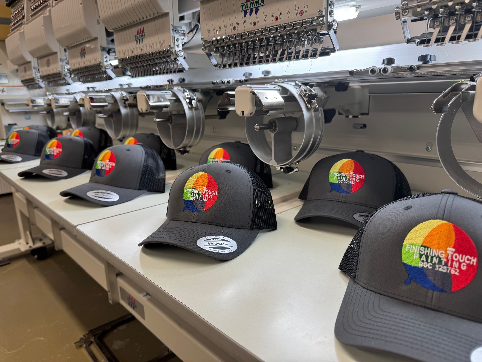 Gray trucker hats with colorful logo embroidered on the front, lined up under industrial embroidery machines.