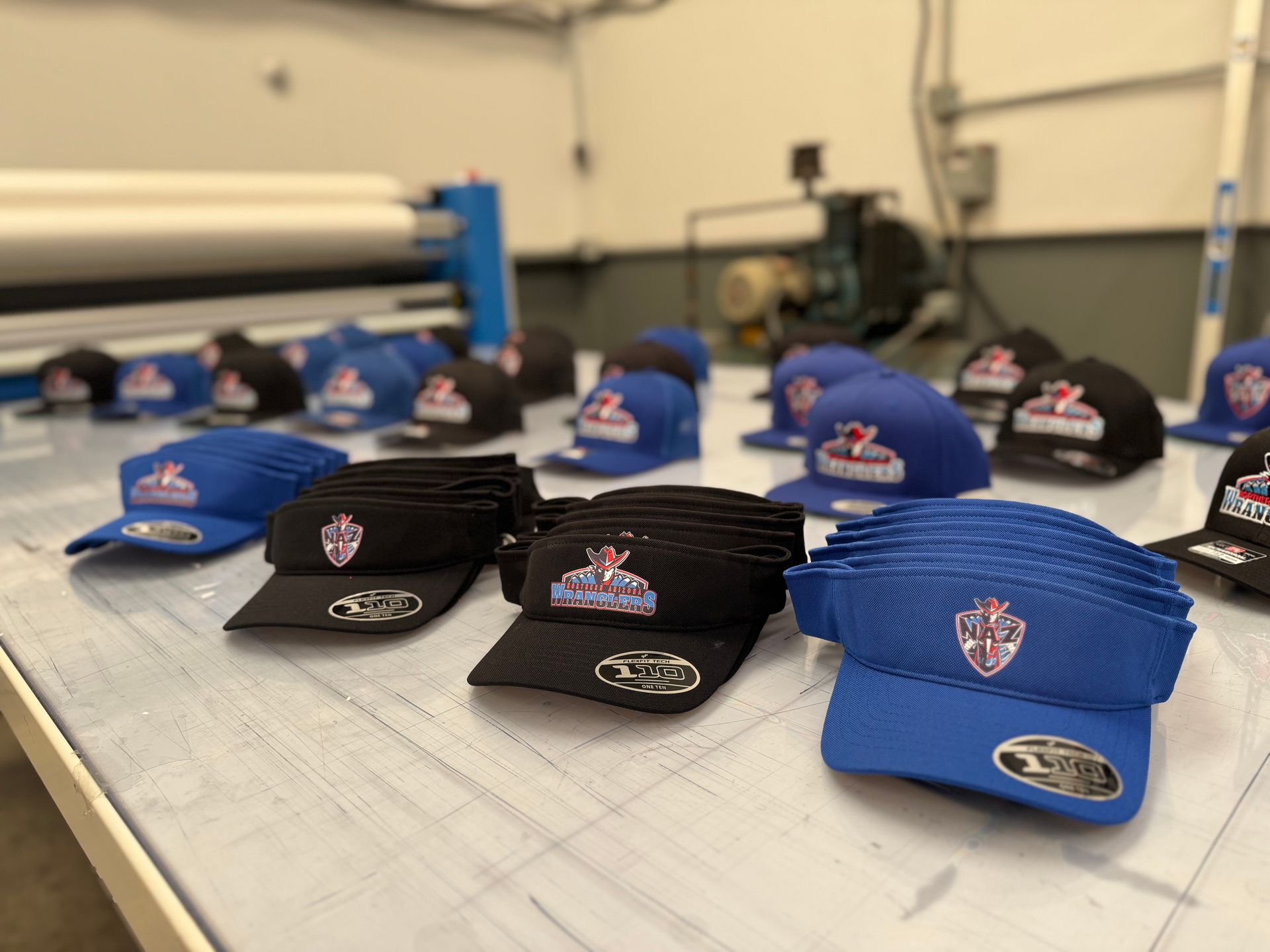 Hats with logos on a table, various colors (blue, black). Background shows machinery in a factory setting.