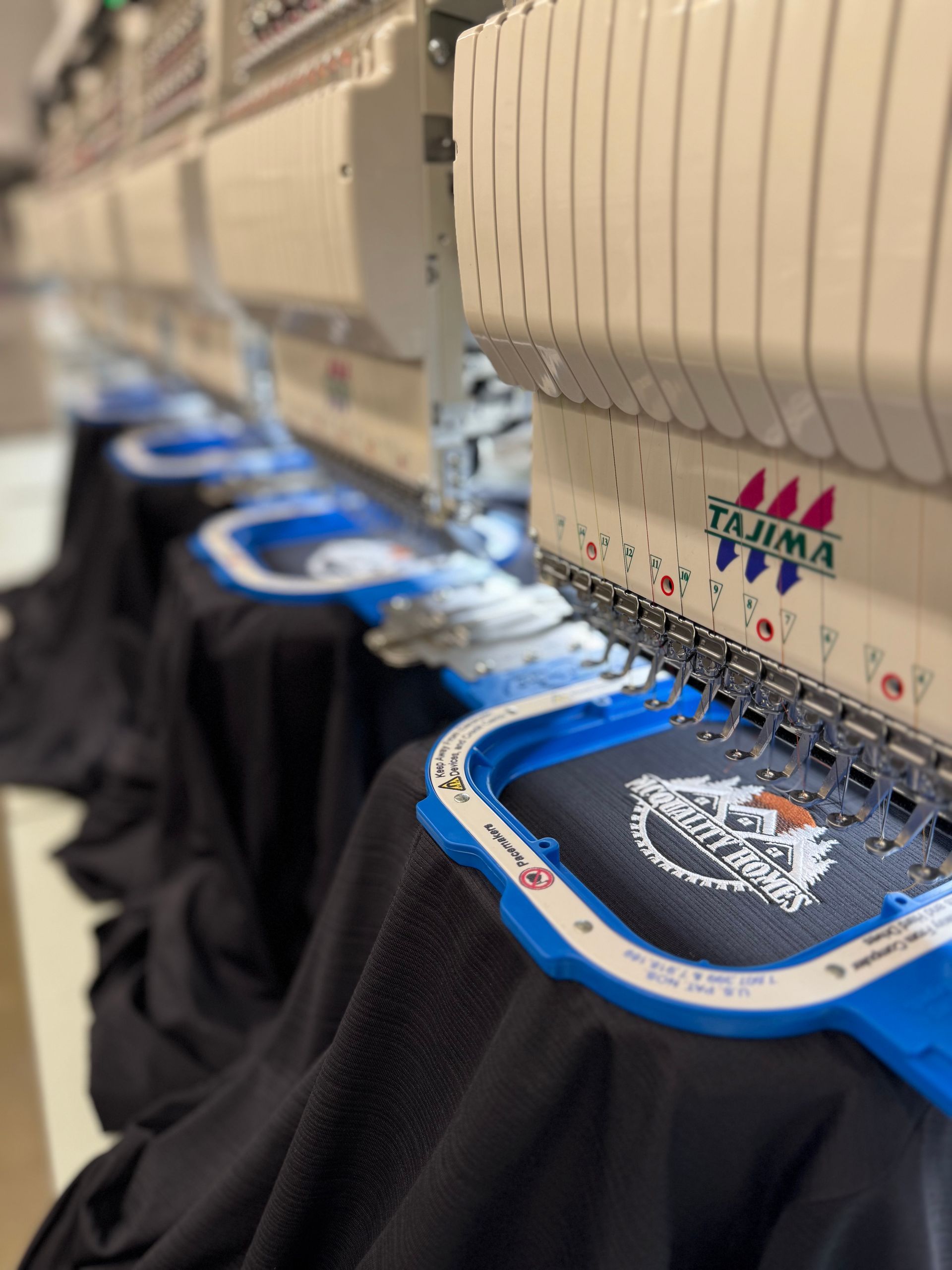 Row of Tajima embroidery machines stitching designs onto black t-shirts.