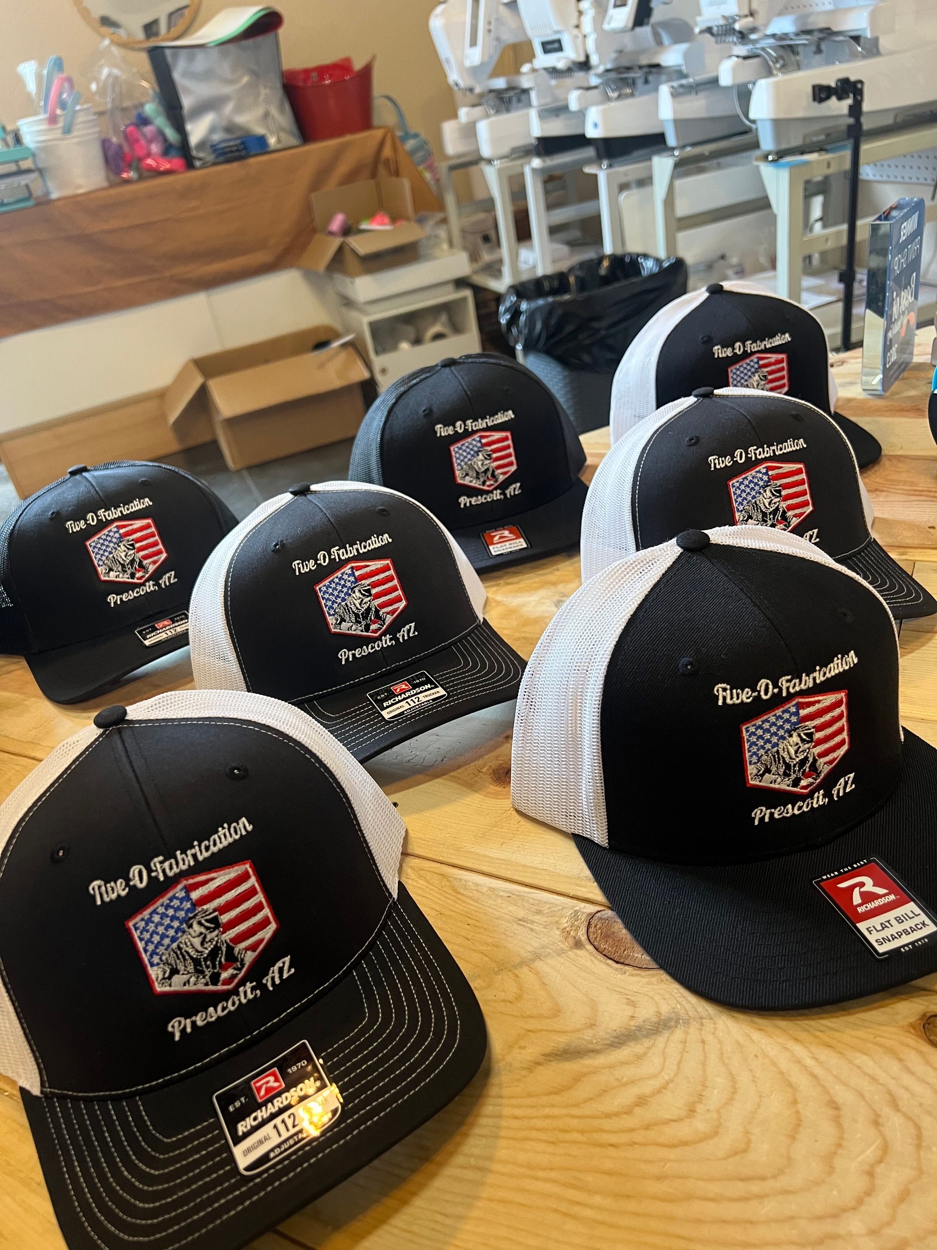 Black and white trucker hats with embroidered patriotic designs on a wooden table. Embroidery machines in the background.