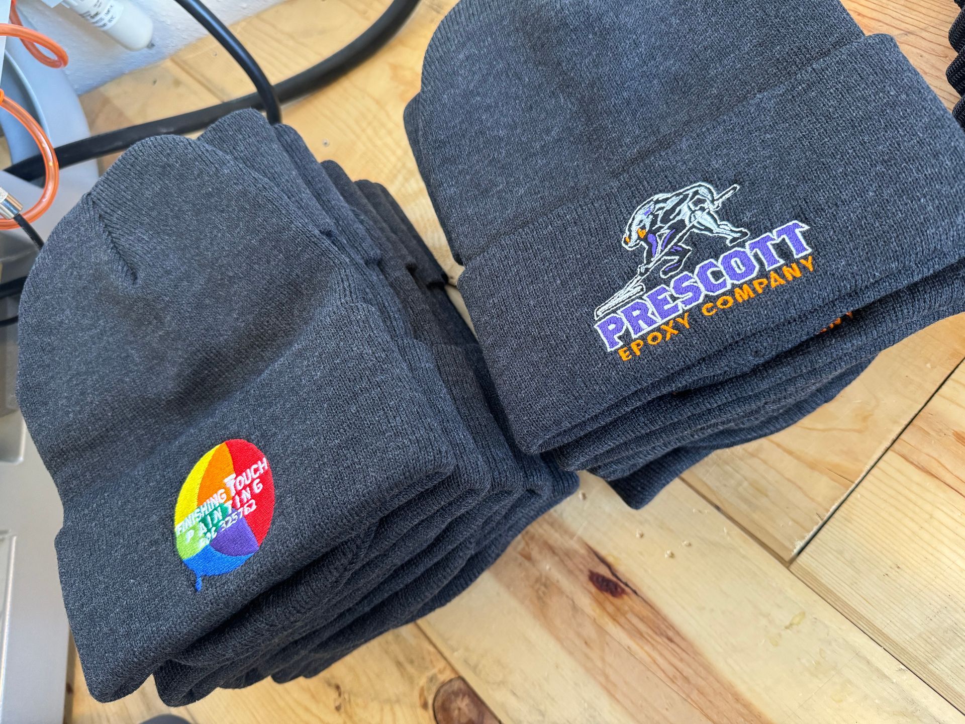 Stacks of dark gray beanies, embroidered with colorful logos: one rainbow and one 