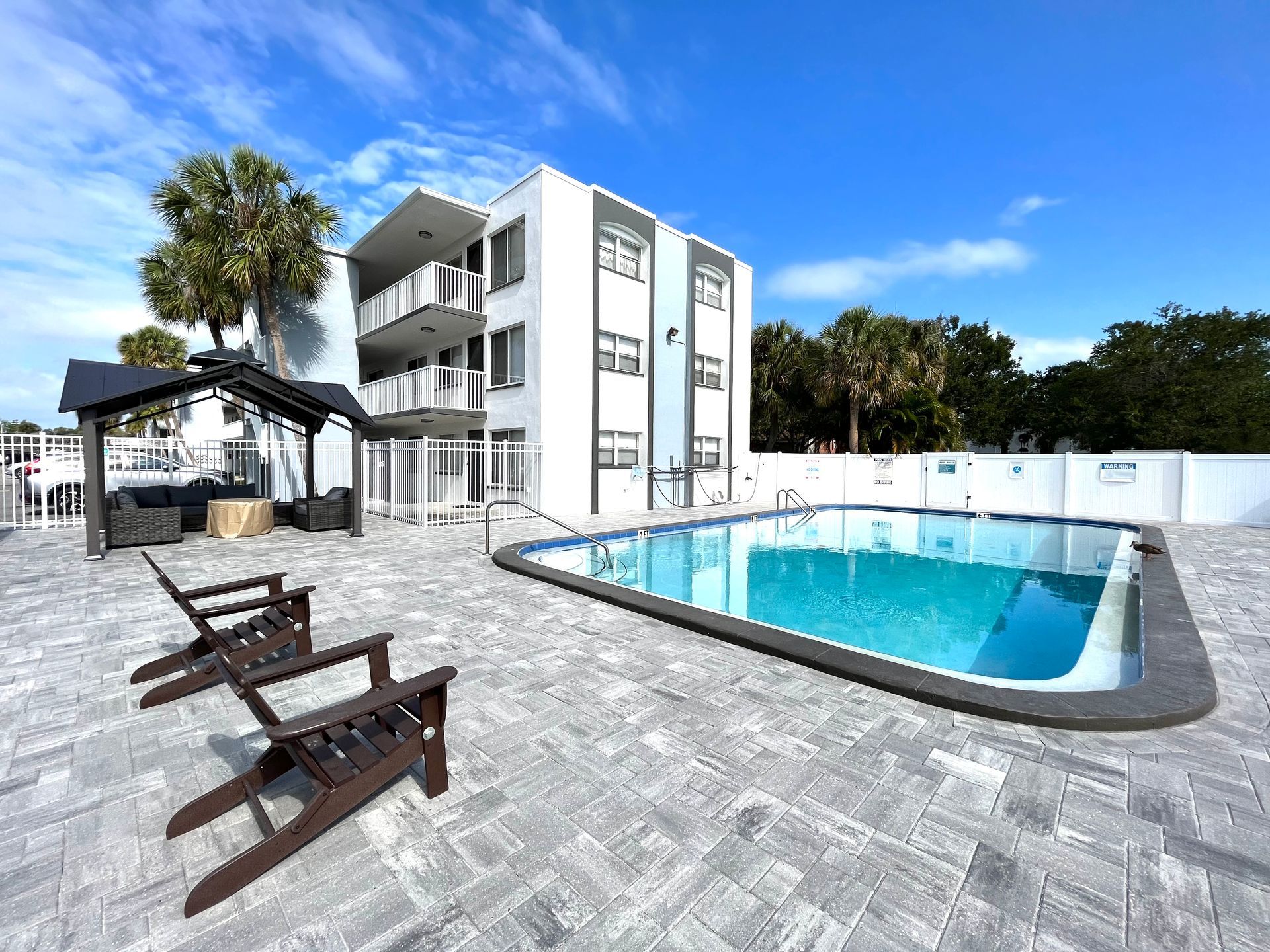Pool area with a building, lounge chairs, and a shaded seating area under a bright blue sky.