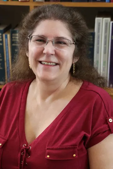 Smiling woman in a red blouse with glasses, seated indoors in front of bookshelves.