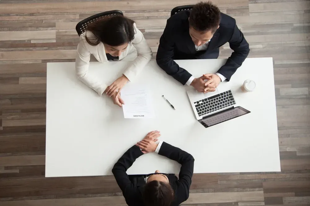Three people in a meeting around a white table with a laptop, papers, and a pen in a modern office.