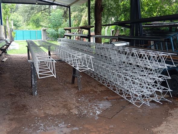 Grandes estructuras de alambre de metal blanco apiladas en un taller abierto bajo un techo, con mesas y árboles cerca.