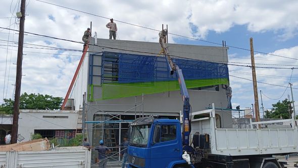 Obreros de la construcción en la fachada de un edificio, con un camión azul y una grúa bajo un cielo nublado.