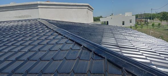 Paneles solares azules en un tejado bajo un cielo despejado, con edificios cercanos y vegetación al fondo.